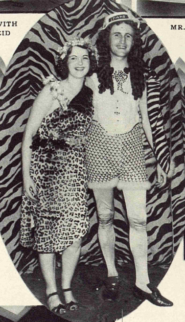 Rosie standing on the left wearing a leopard print dress and sandals, with a flower garland in her hair. Mr Ogilvie Smith's outfit includes a wig of long, dark curly hair, a boater hat, printed shorts with fur lined cuffs, silk tights and pointed shoes.