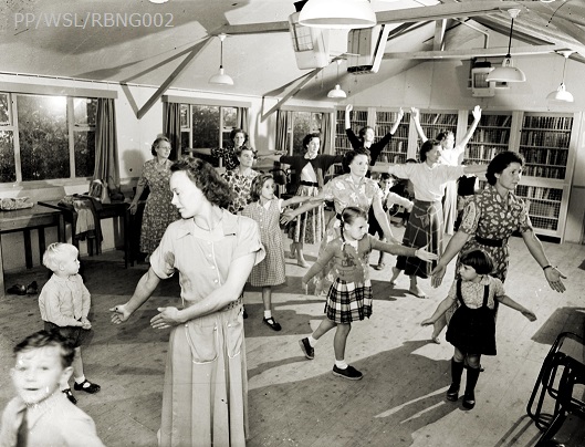 Black and white photograph of the Young Wives Club in Langley Green. The women are dancing with their children in a community hall.