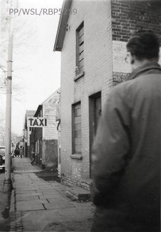 Black and white photograph of the back  of a coat-clad man walking down a run-down street in Crawley.
